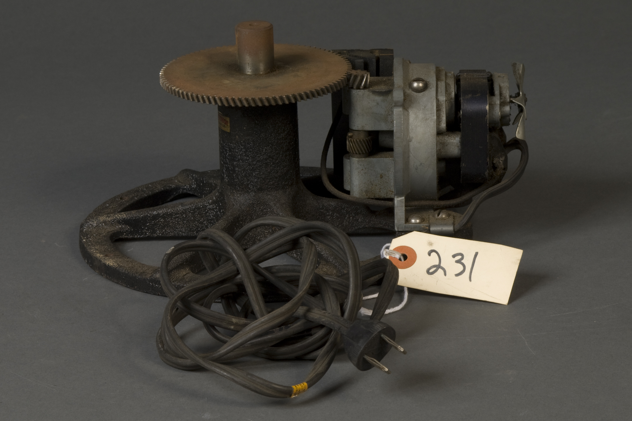geared mechanism with an electric plug. Attached to the plug's wire is a tag that reads "231." The device is geared to another smaller gear on a device with what looks like a flywheel coming from the front of it. 