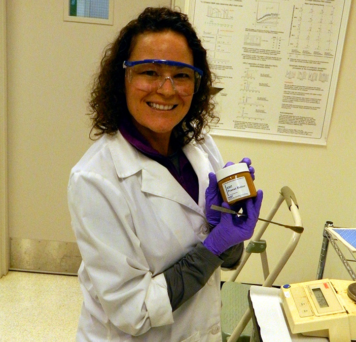 NIST chemist Carolyn Burdette in a white lab coat and wearing goggles holding up a jar of  SRM 2387, Peanut Butter