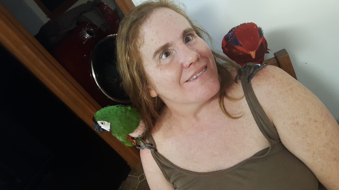 Mrs. Hidle and her birds, Ned, a green parrot, sits on her right shoulder, while Mimi, a red parrot, sits on her left 