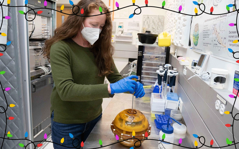 A woman wearing a mask stands over a yellow sphere at a lab bench. 