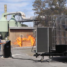 A photo shows an outdoor experimental setup with a huge fan blowing fire toward a shed. 