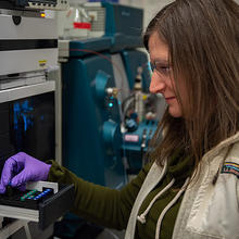 Melissa Phillips wears safety glasses and gloves in the lab as she places small vials into a rack in a large machine.