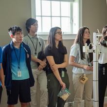Half a dozen high school students stand listening to a presentation in a room with scientific equipment.
