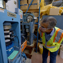 A researcher in safety gear leans over to examine a concrete core sample placed inside a blue metal testing machine.