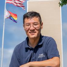 Dr Raymond Sheh in front of Building 101 and main flagpole, flying the US and Progressive Pride flags, at the main NIST campus in Gaithersburg. 