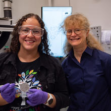 Diana Ortiz-Montalvo, wearing safety glasses and holding a small circular filter with tweezers, poses with Abigail Lindstrom in the lab.