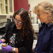 Diana Ortiz-Montalvo looks into a small container in the lab while Abigail Lindstrom stands behind her. 