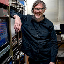 Andrew Novick poses leaning on a rack of servers and other electrical equipment.