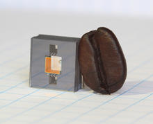 Square gray cube with two square holes in it and a gold square in between the holes, and a dark brown coffee bean leaning against the cube.
