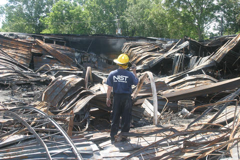 Man in NIST fire research t-shirt and hard hat standing among wreckage