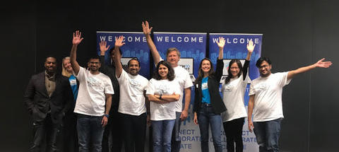 This image shows a group of challenge participants, most in white t-shirts, standing in front of a "Welcome" sign for the Tech to Protect Challenge.