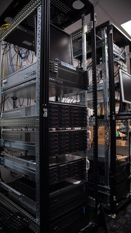Tall black racks hold computer servers, with wires hanging down around them. 