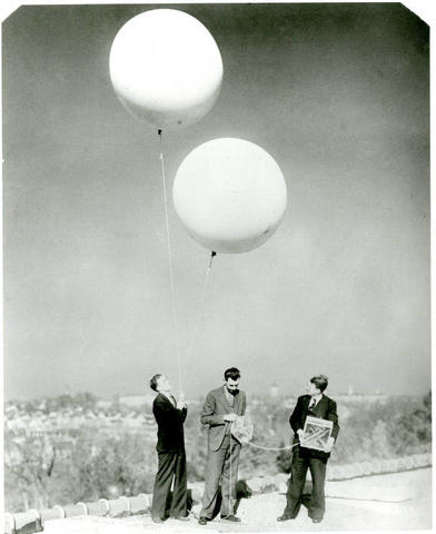 Historic photo shows three men in suits standing on the roof of a building, preparing two large white balloons with attached instruments.