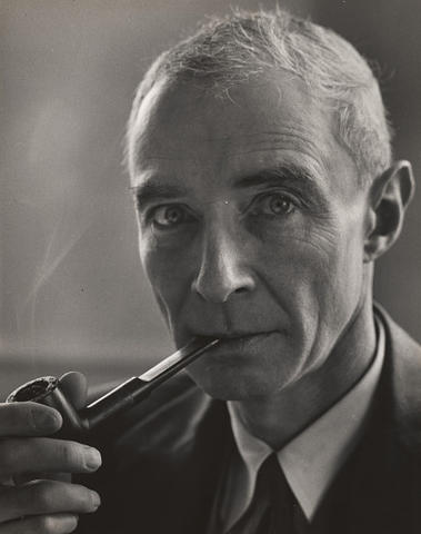 J. Robert Oppenheimer, in a black and white photo, peers into the camera while holding a pipe in his mouth. Smoke billows out from the pipe. 