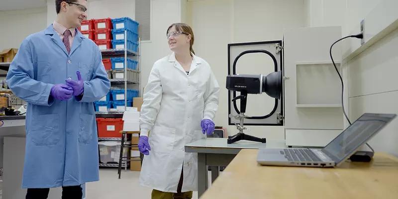 Ryan Falkenstein-Smith and Amy Mensch, both wearing lab coats and gloves, stand in the lab next to a black camera-like device on a table. 