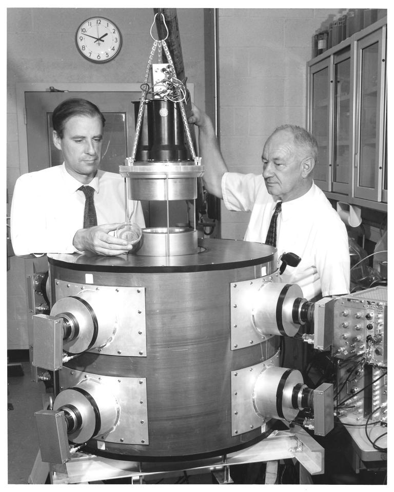 Two men stand on either side of a large cylindrical device.