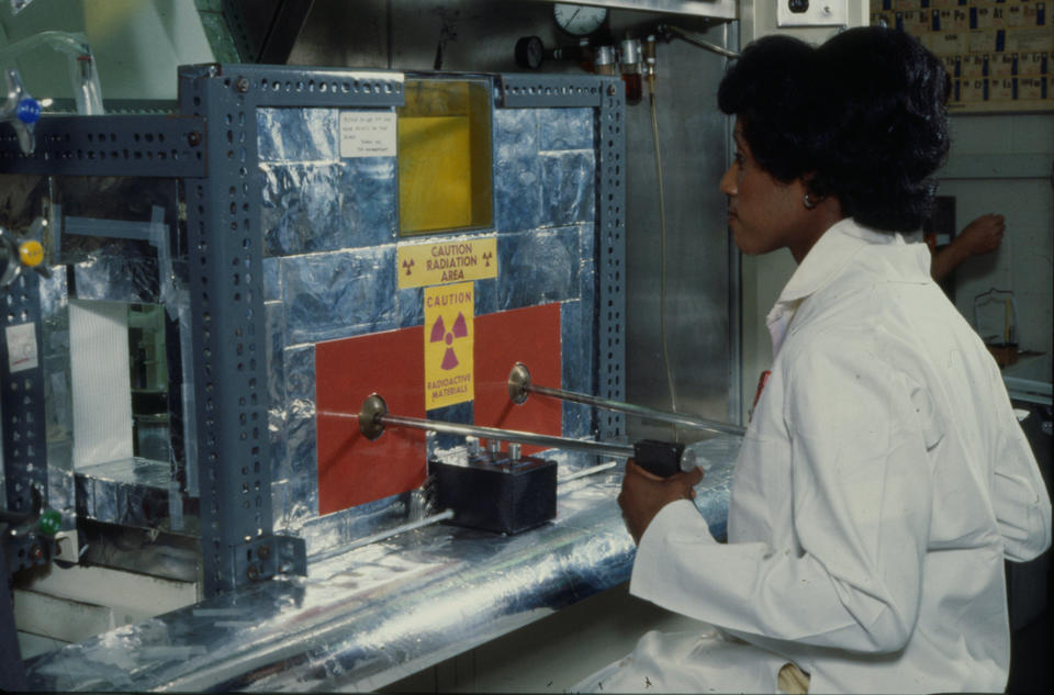 A woman works at a metal machine with a radioactivity warning sticker