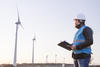 A man in a hard hard is holding a clipboard and looking towards a large wind turbine. 