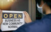 Person wearing a mask posting a sign on a glass door that says Open Business As New Community Normal