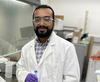 George Caceres wears safety glasses and a lab coat as he poses for a head shot in the lab. 