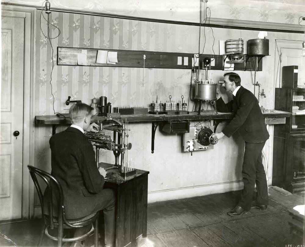 Man sits at a desk on the left while a man on the right stands looking into a device