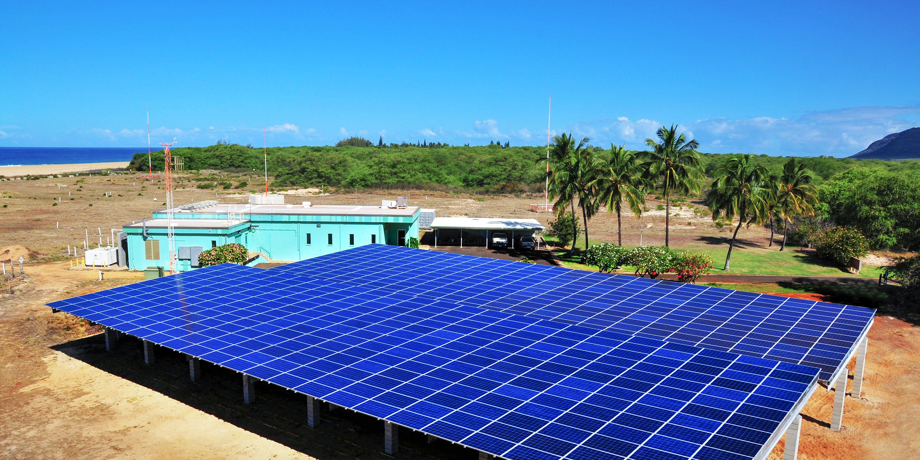 view of an array of blue solar panels with a turquoise building behind them on a barren patch of soil. The beach and ocean are in the distant background. 