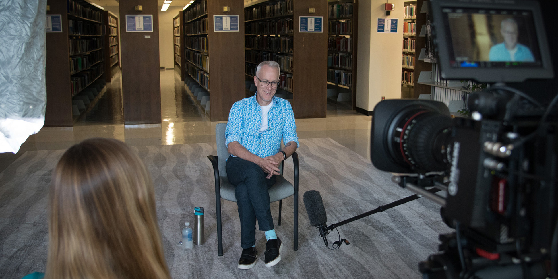 Jon Pratt, wearing a blue and white patterned shirt, sitting in a library in a chair with legs crossed, smiling to himself, with a film crew mostly out of frame in the foreground