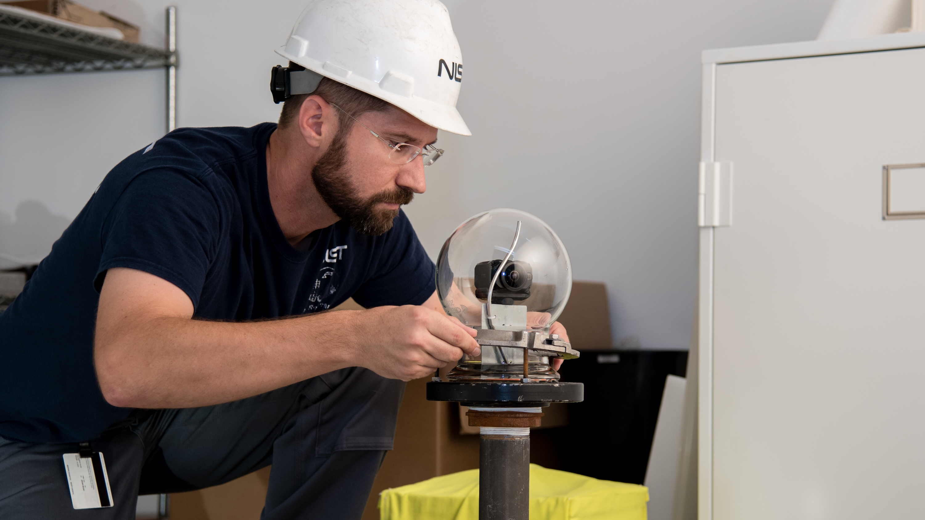 man with a beard, safety glasses, hard hat and blue shirt places a glass bubble over the top of a camera mounted on a steel pole inside the test room