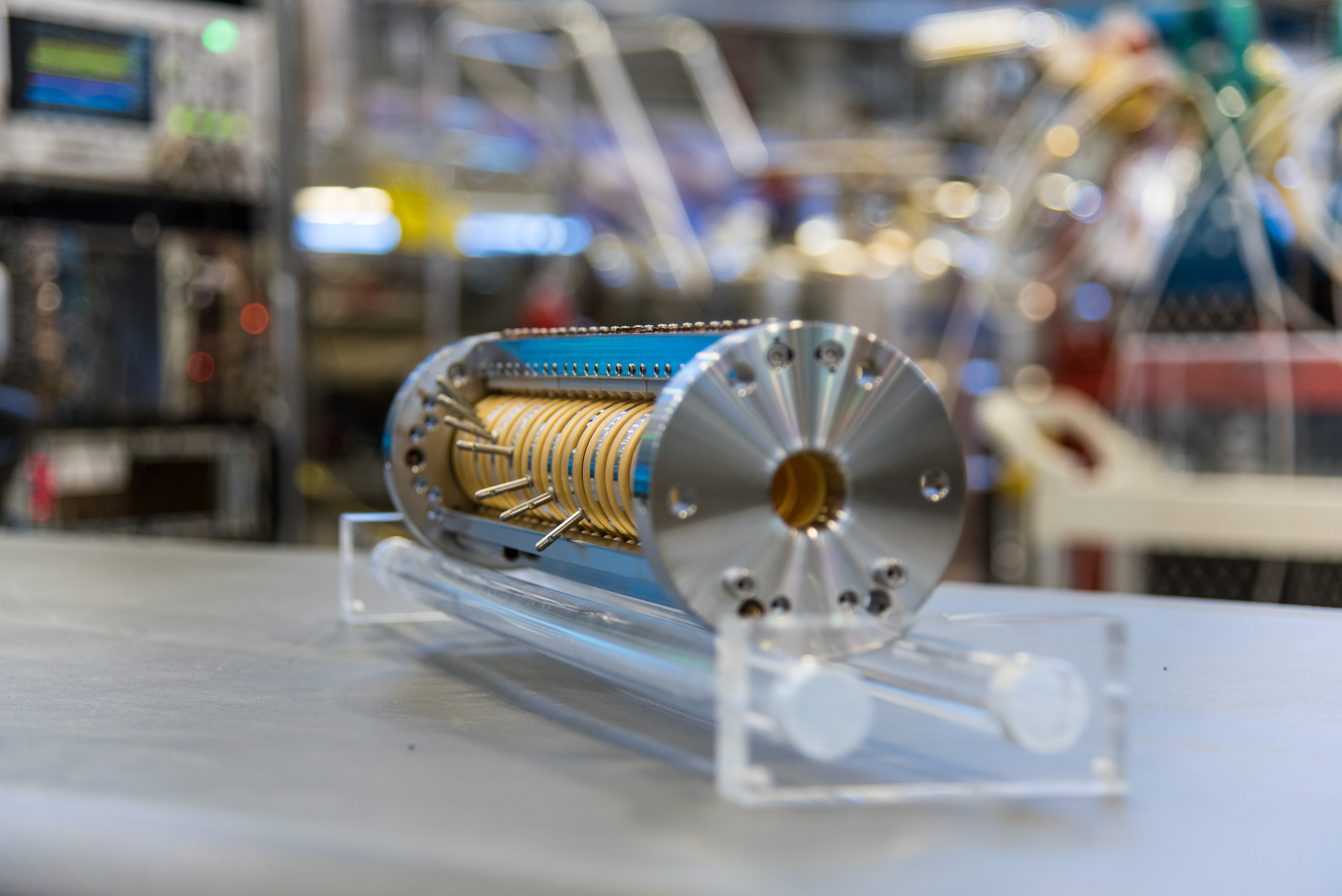 cylindrical rod of metal-banded disks capped by aluminum donuts resting on a metal table. A band of blue circuitry runs along the top.  