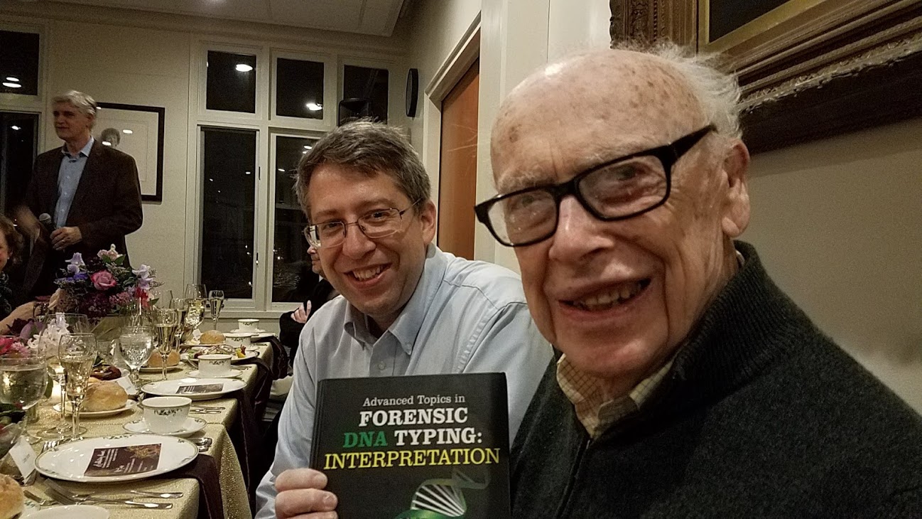 John Butler and James Watson sitting side by side. Watson is holding a copy of Butler's book up for the camera. 