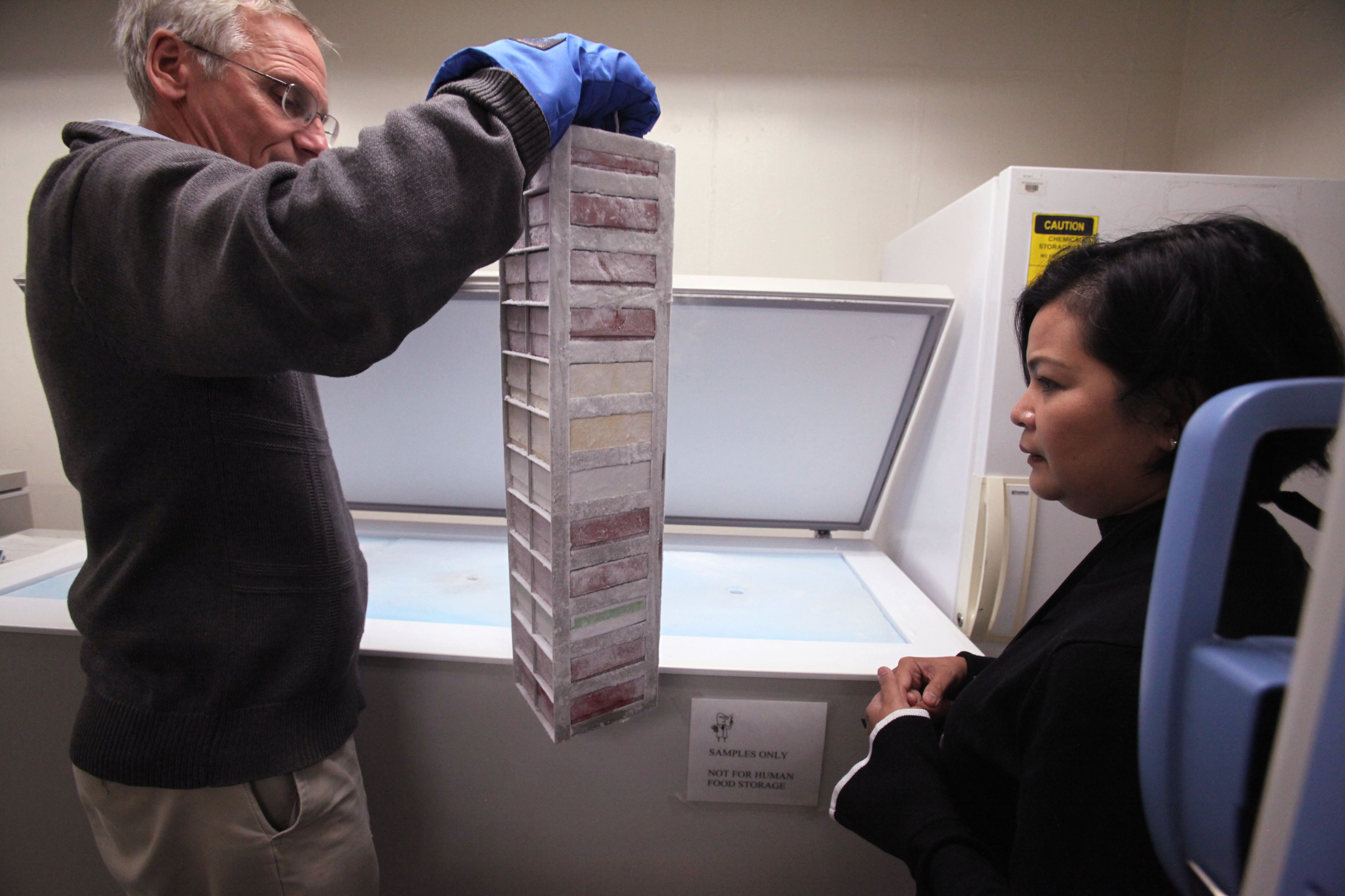 Man stands in front of a freezer holding a stack of milk vials. NIST scientist stands next to him.