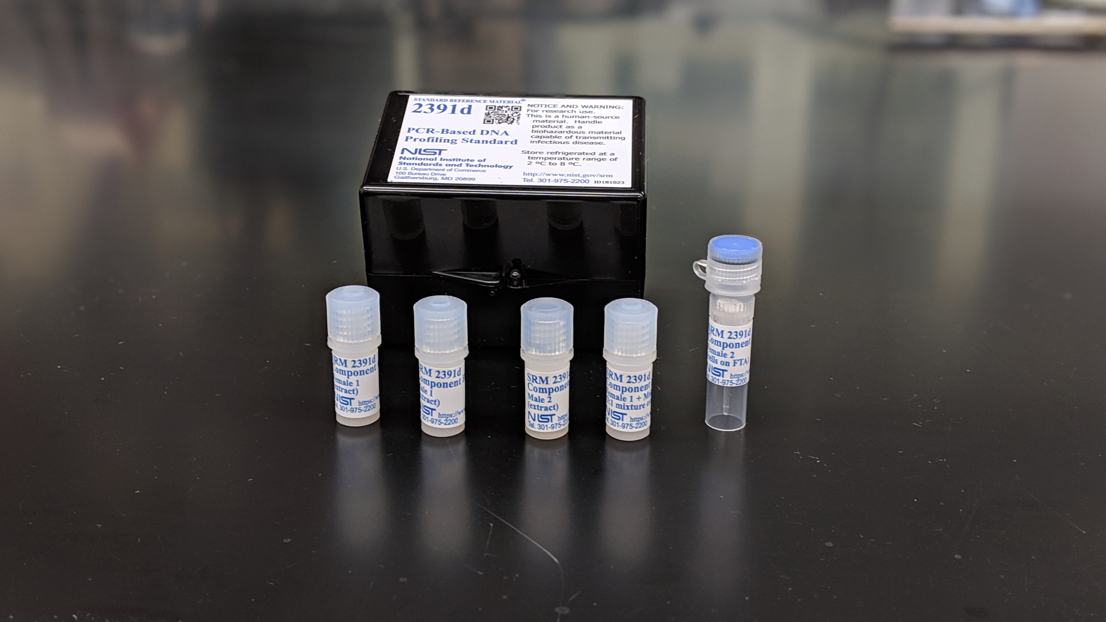 portrait of five plastic vials on a table. There is a black box behind them that reads "2391d PCR-Based DNA profiling standard."