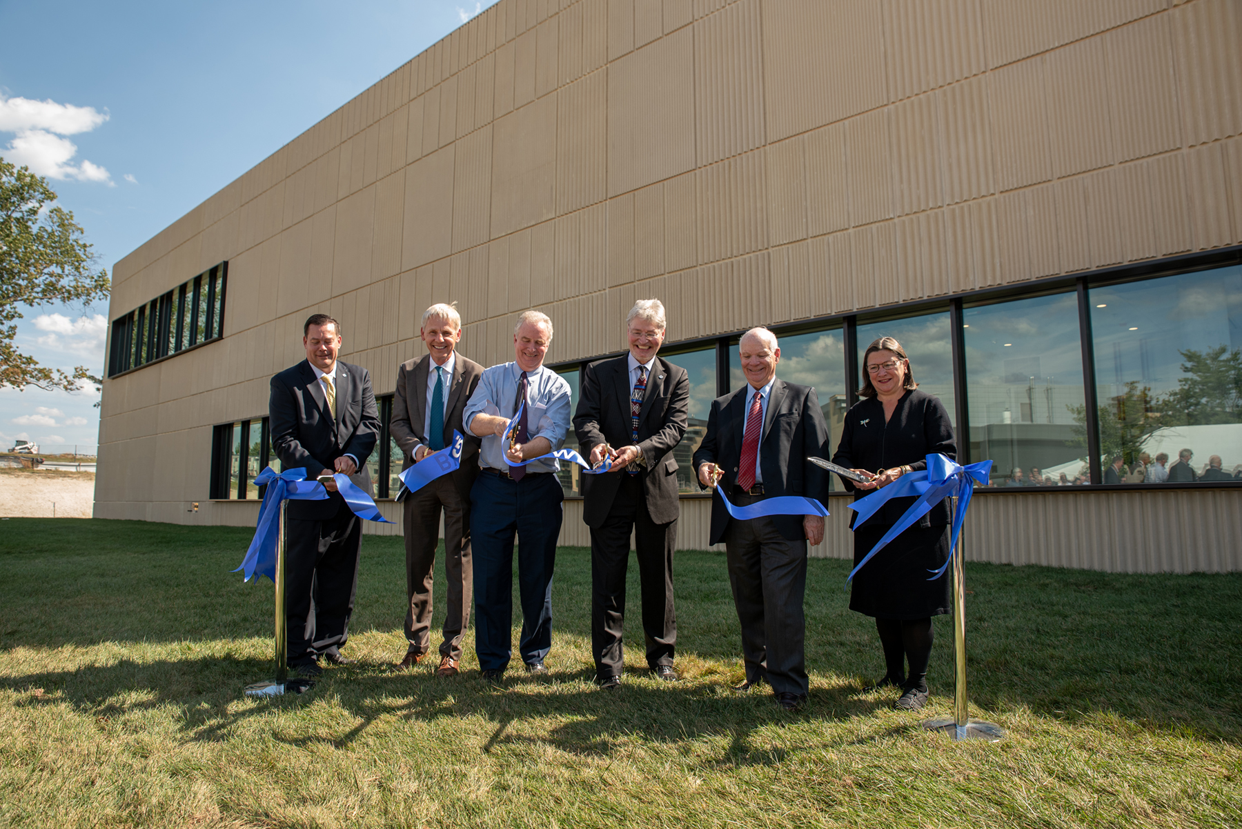 Six people stand in front of a tan building, cutting a blue ribbon