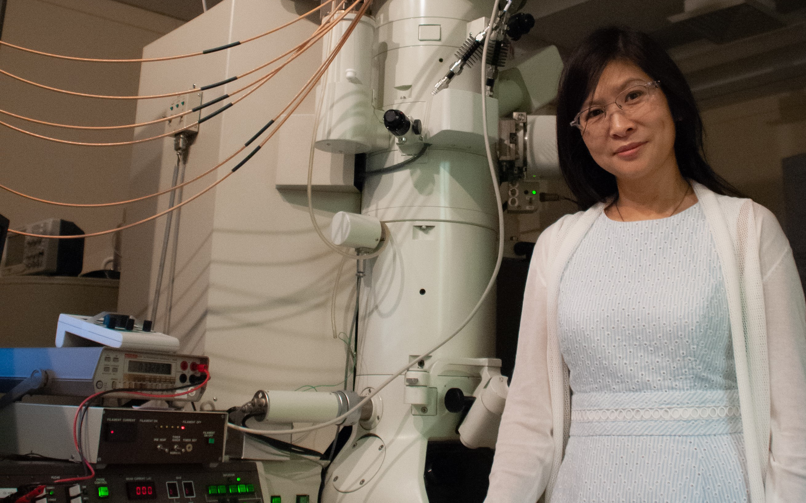 woman in a blue dress and white sweater stands next to an electron microscope