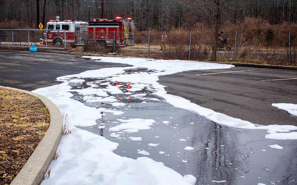 A parking lot with a large puddle of aqueous firefighting foam. A firetruck is in the background. 