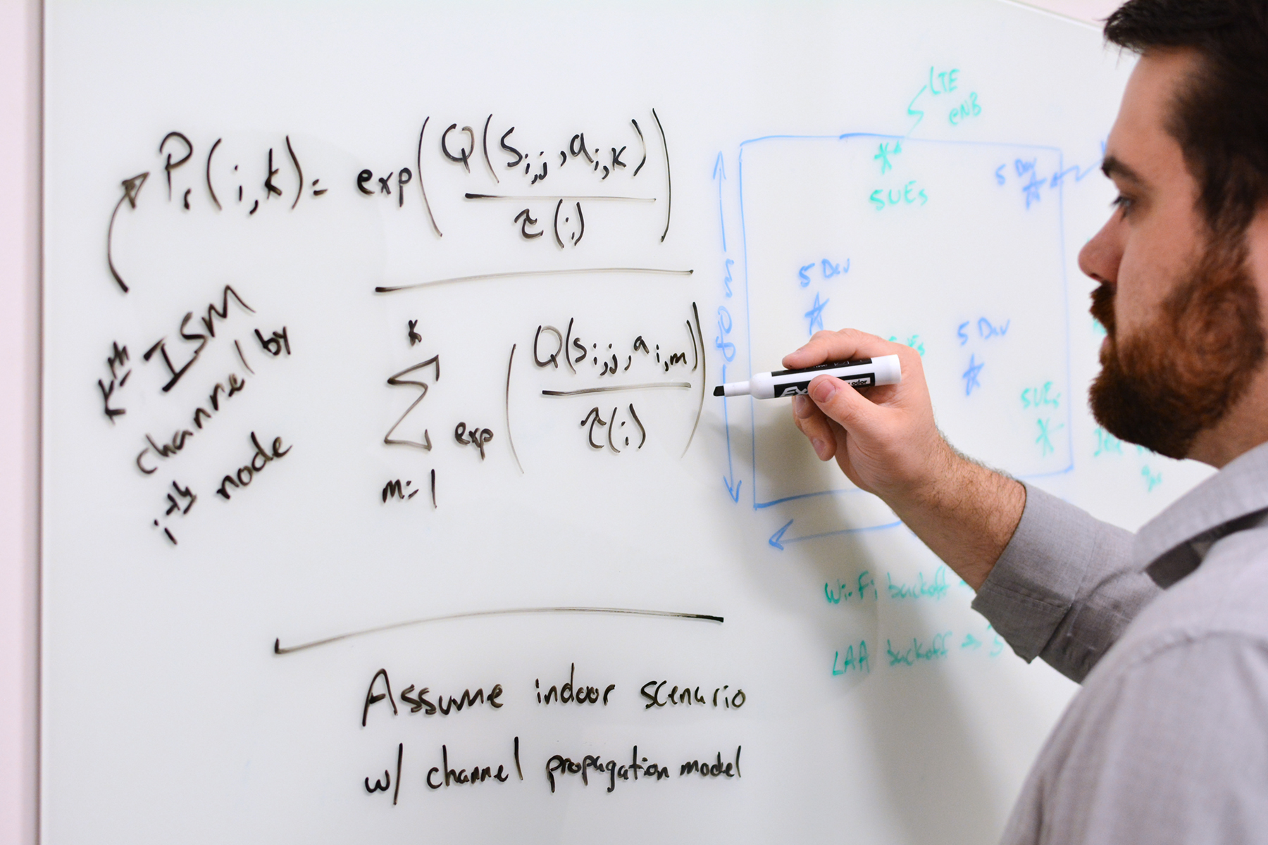 Man with dark hair writing mathematical formulas on a whiteboard on a wall.