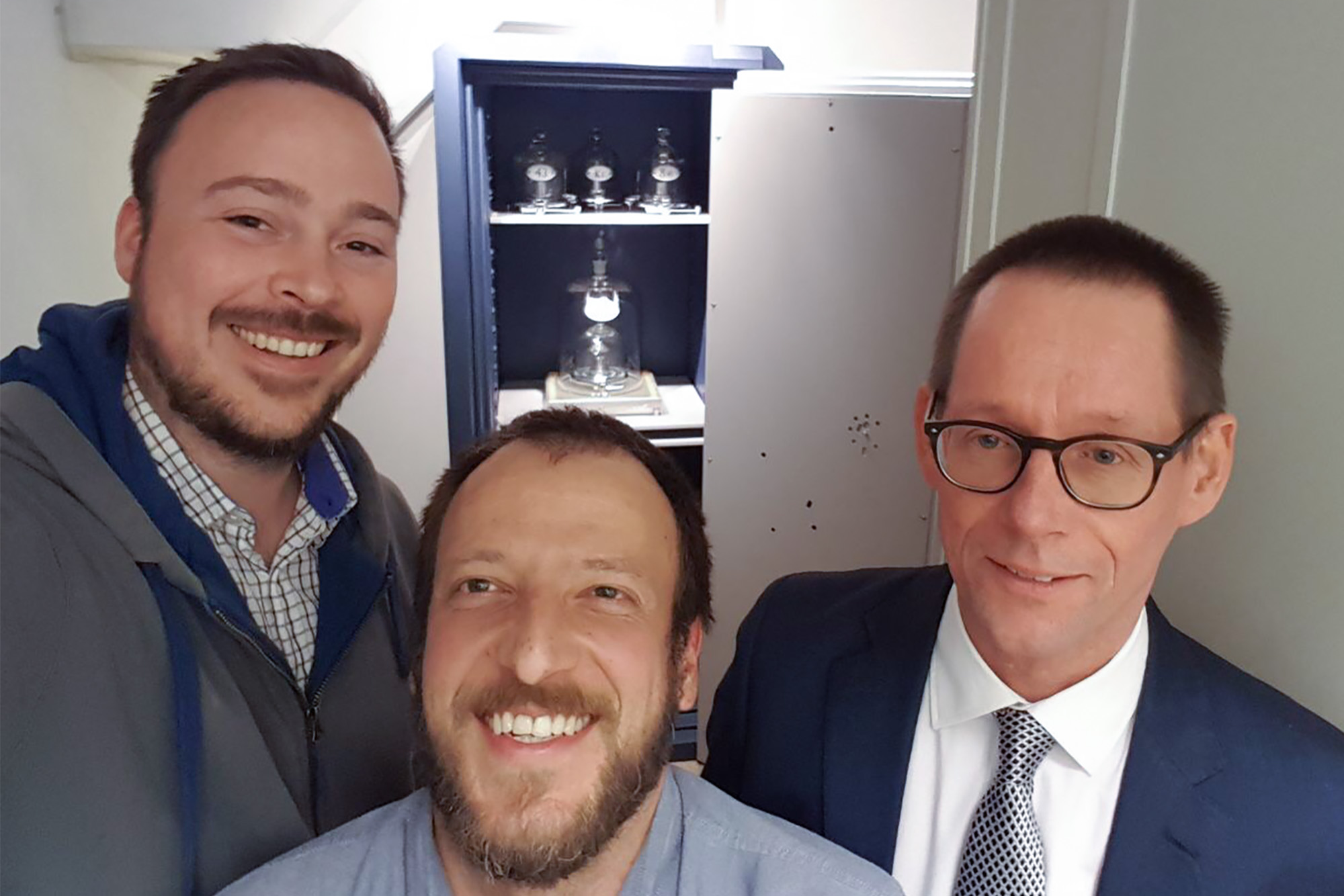 three men take a selfie inside the kilogram vault. The kilogram is visible in a safe behind them. 