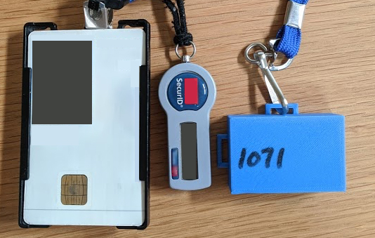 a small, blue 3D-printed box, which is a wearable exposure notification device, next to an RSA remote token and an ID card for scale. The ID card is about the size of a credit card and the device is about one third its size.