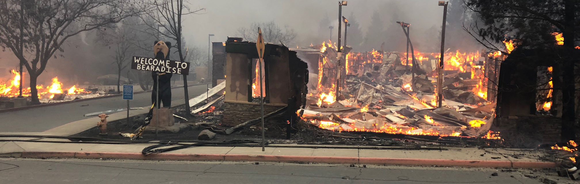 Welcome sign stands next to a burning building.