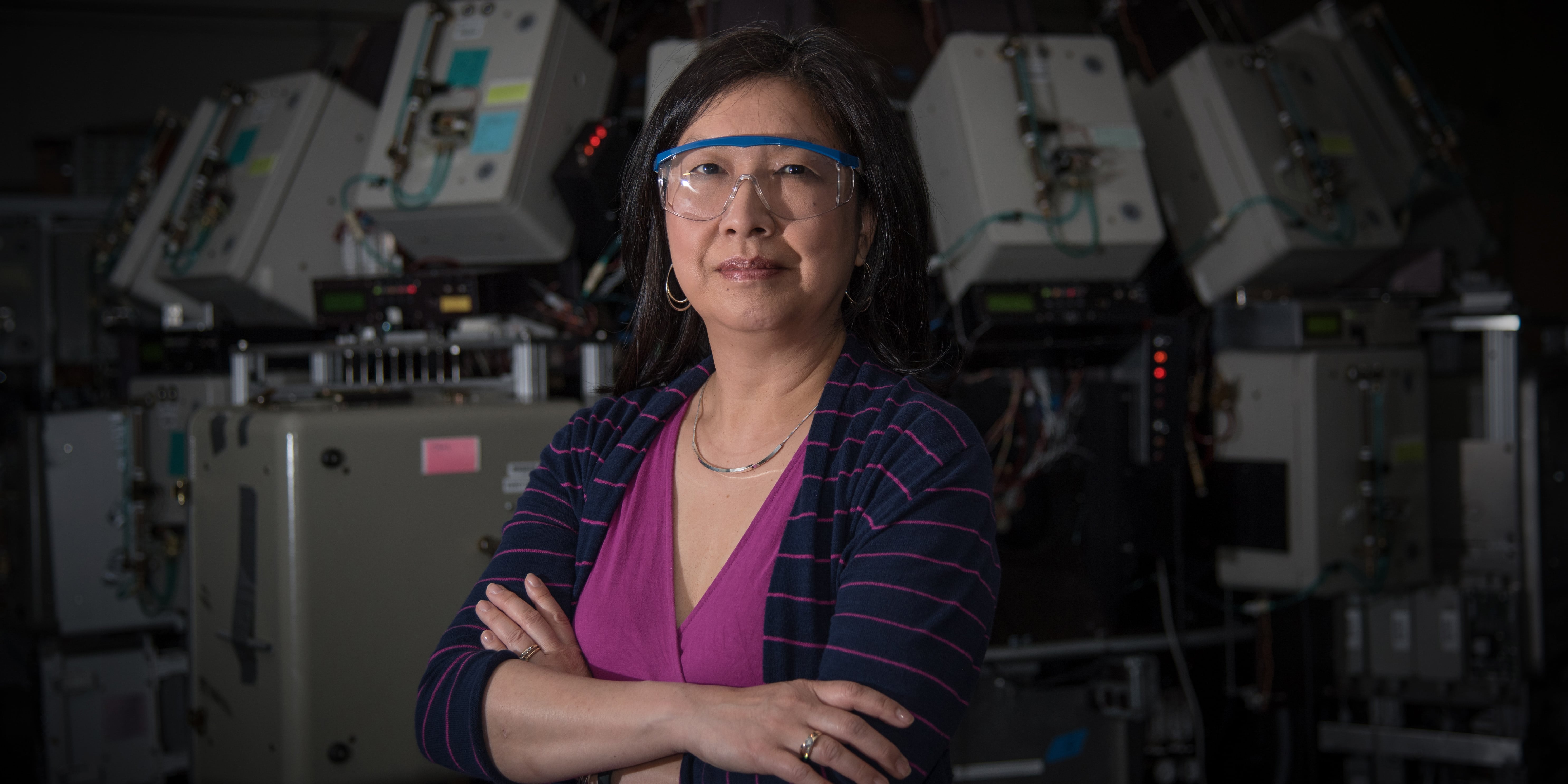Joannie Chin stands with arms crossed, wearing safety goggles, in front of scientific equipment. 