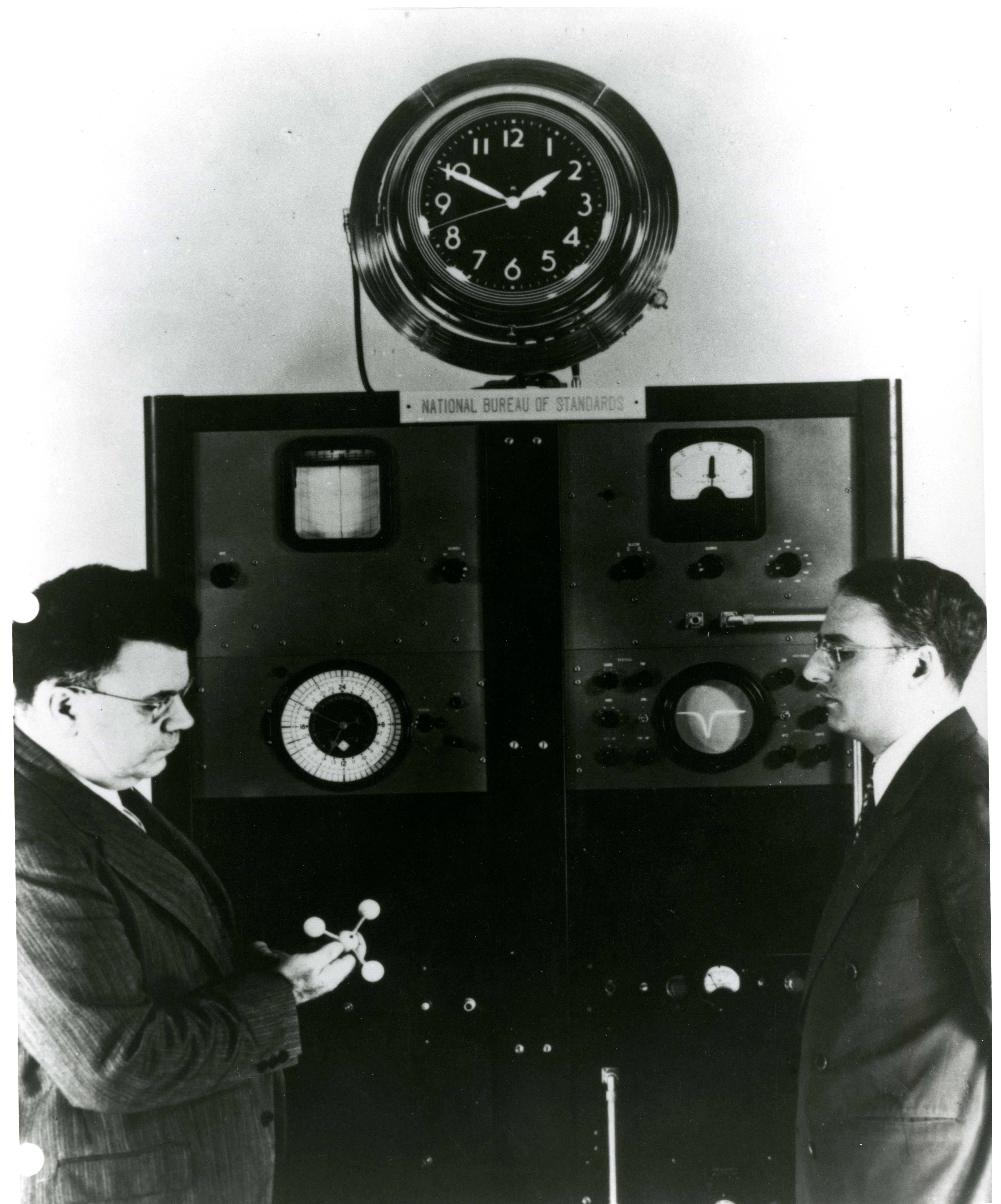 Photograph of NBS director Dr. Edward Condon poses with Harold Lyons, head of the NBS clock program, in front of the first atomic clock.