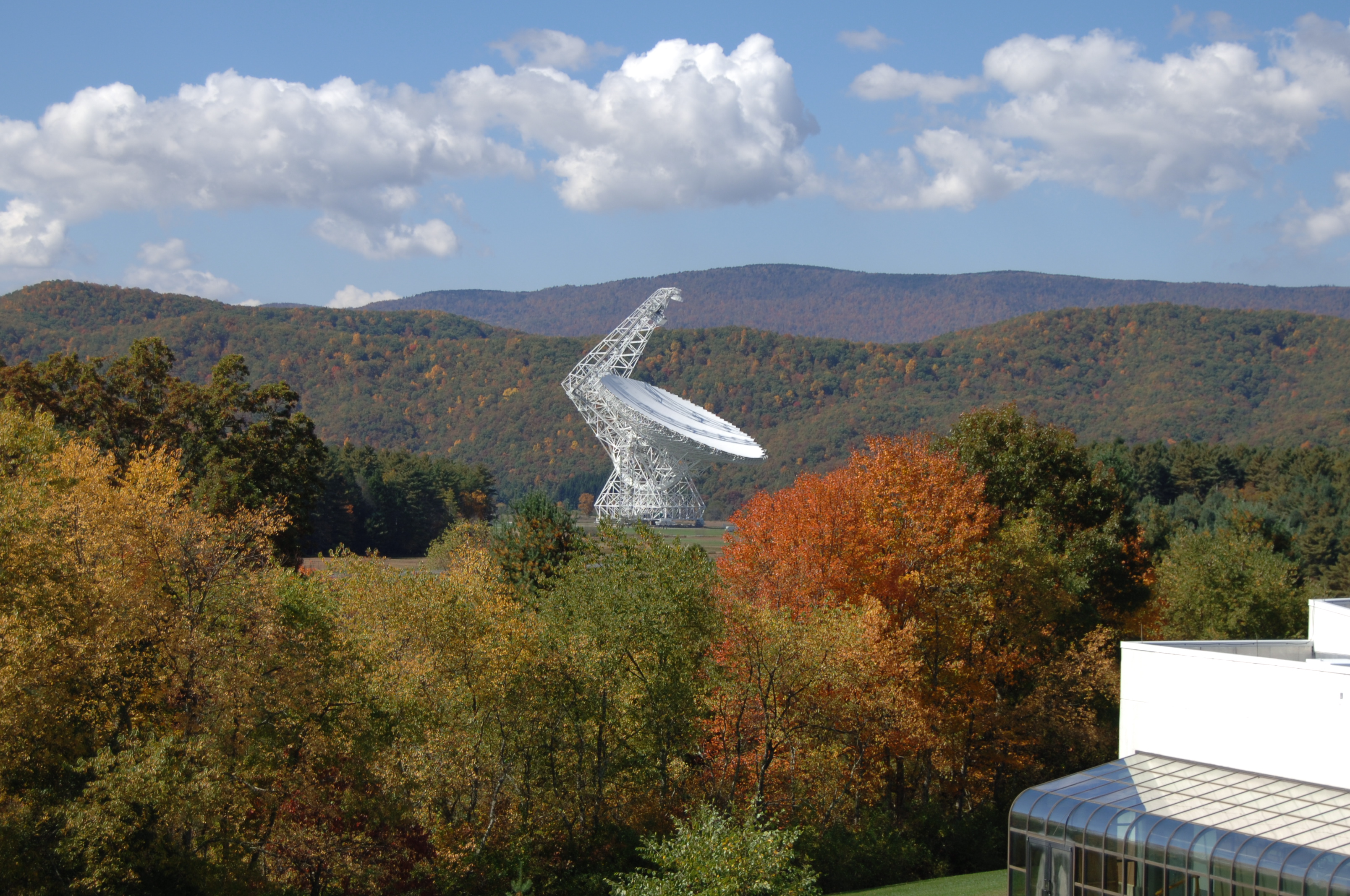 A white telescope dish is surrounded by wooded mountains in autumn colors.