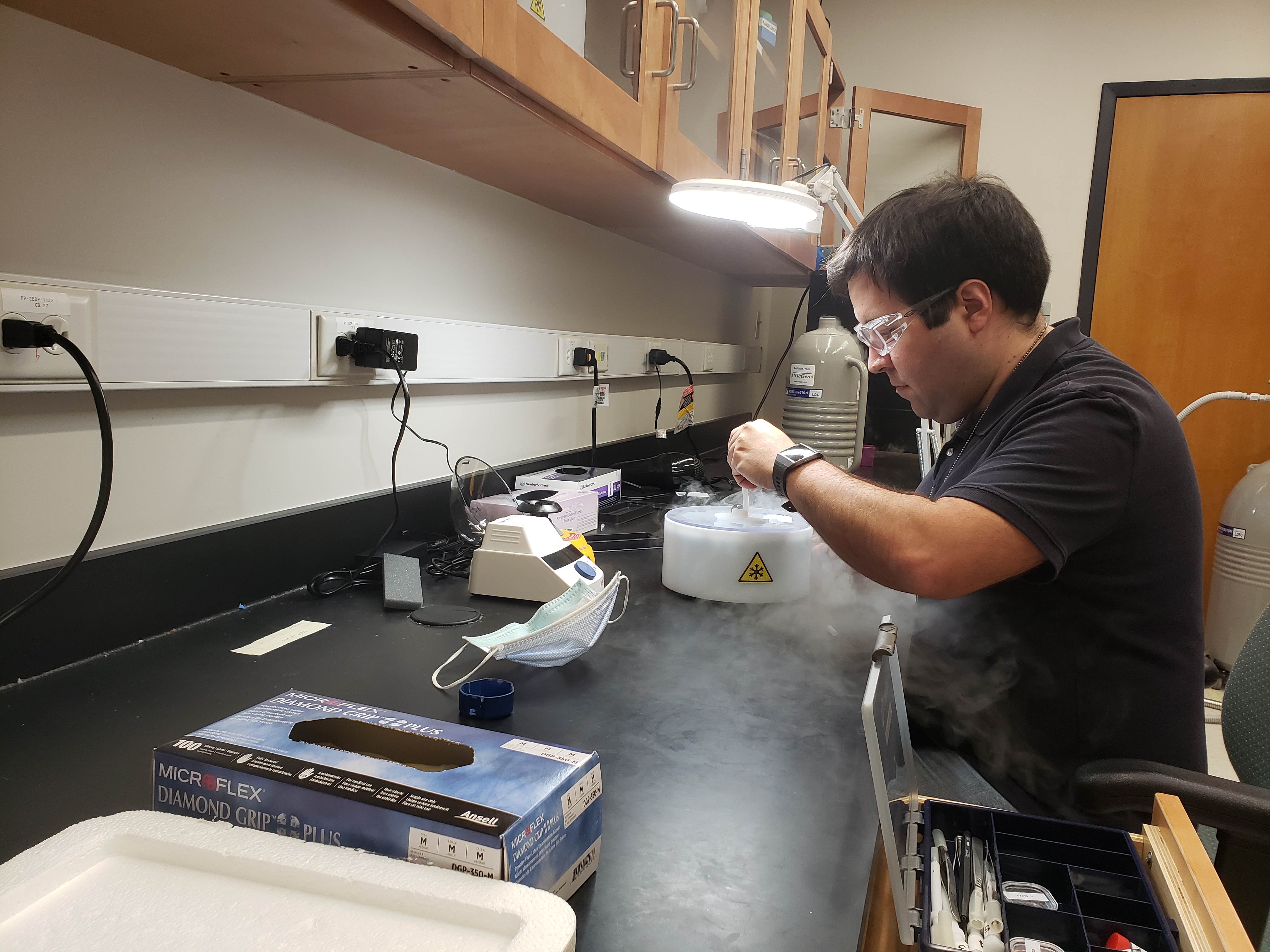 man with safety glasses sitting at a lab bench looking into a container smoking with liquid nitrogen.