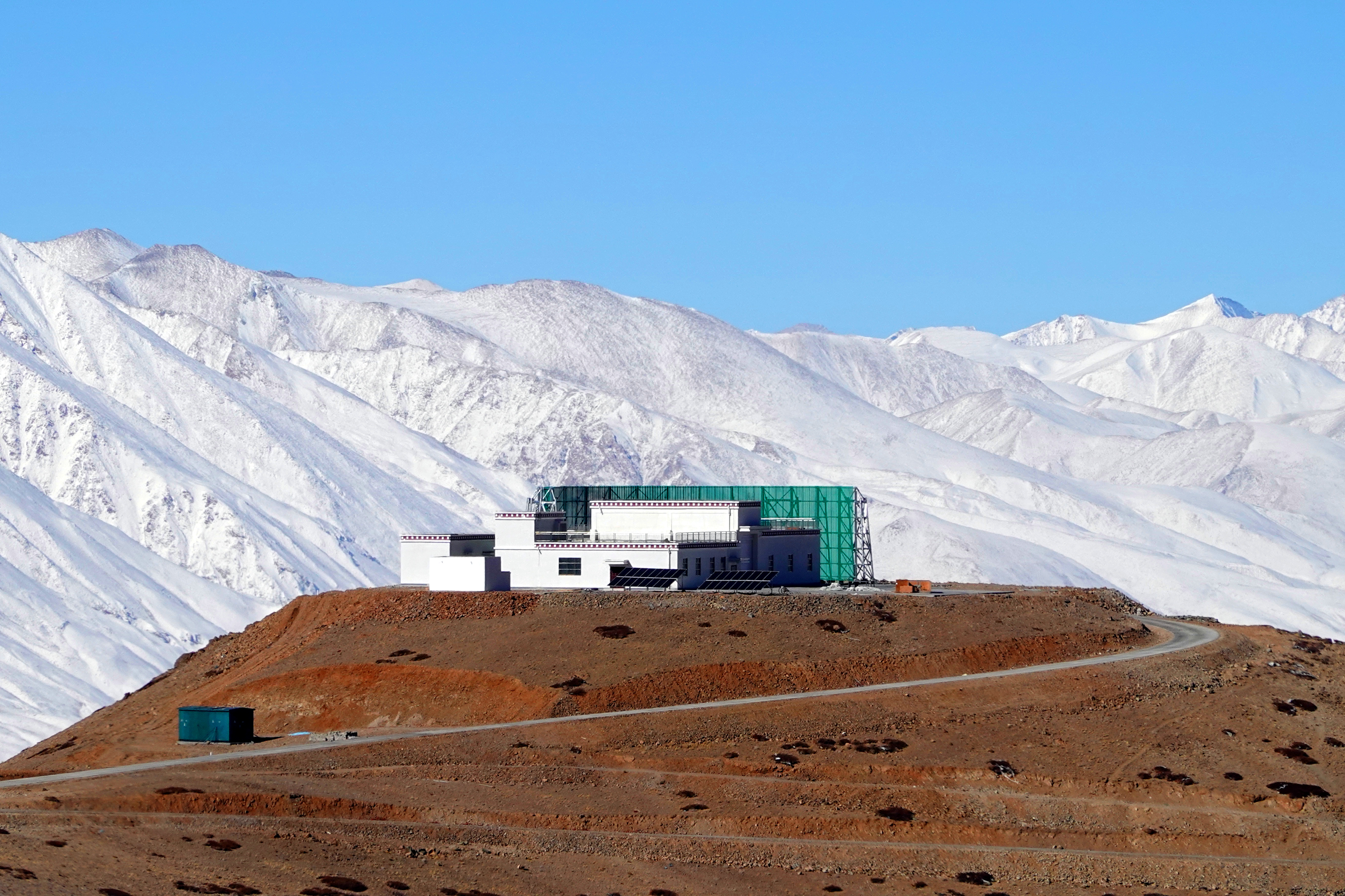 Blocky building sits on dirt hill with snowy mountain and blue sky in background.