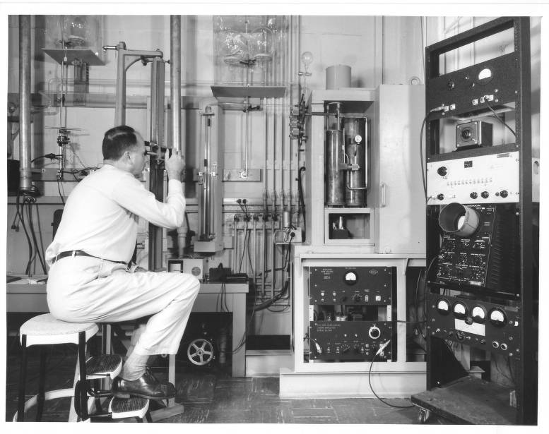Historical photo shows a man dressed in white sitting on a stool working with a wall of scientific equipment.