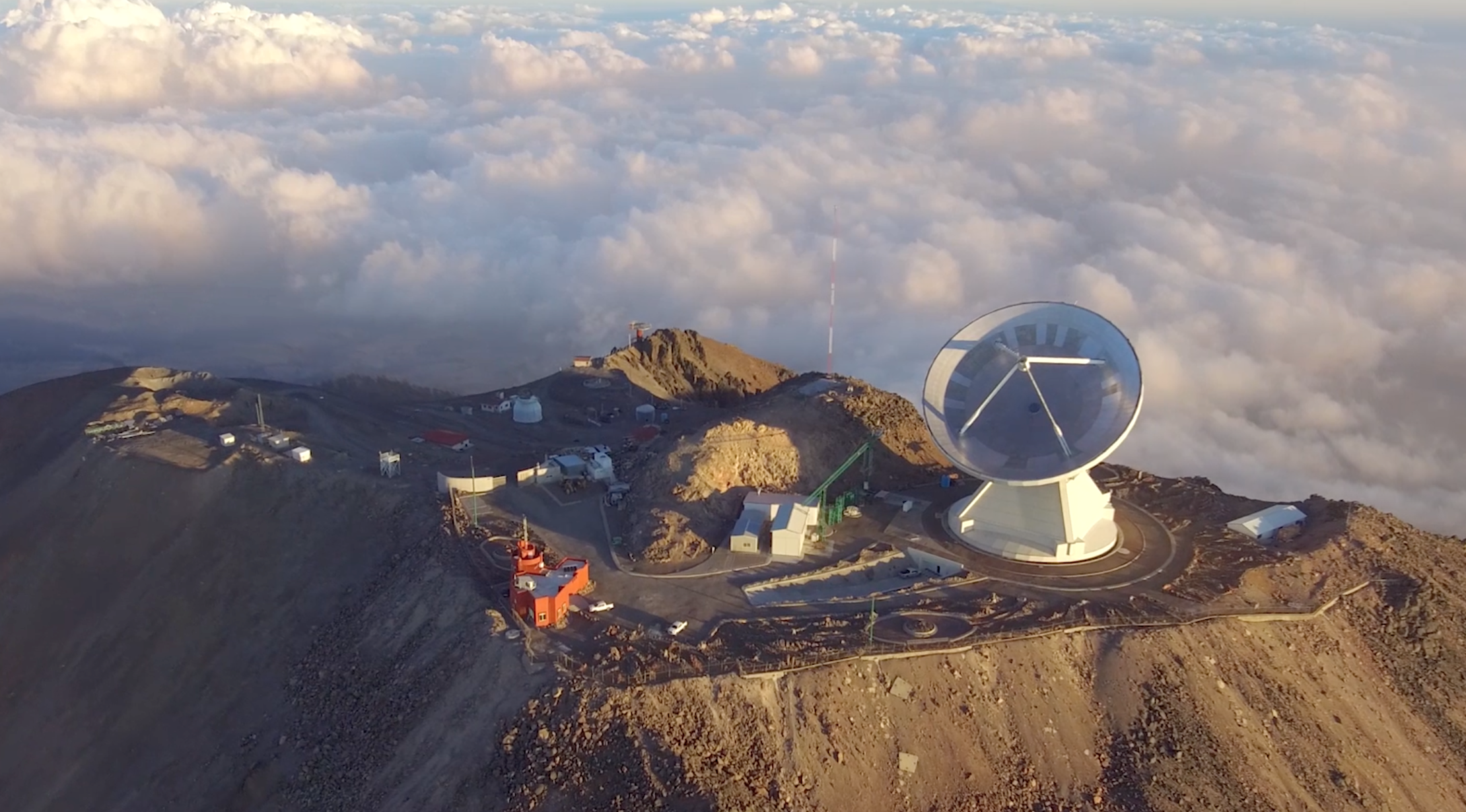 Large dish is on top of a mountain with clouds at the same level. 