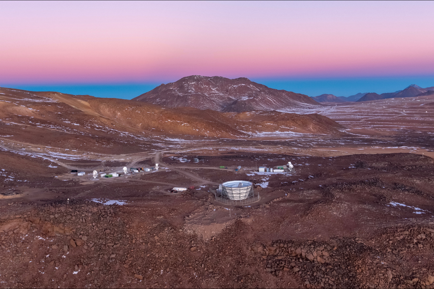 A white telescope dish is set in the desert with mountains and a colorful sky in the background.