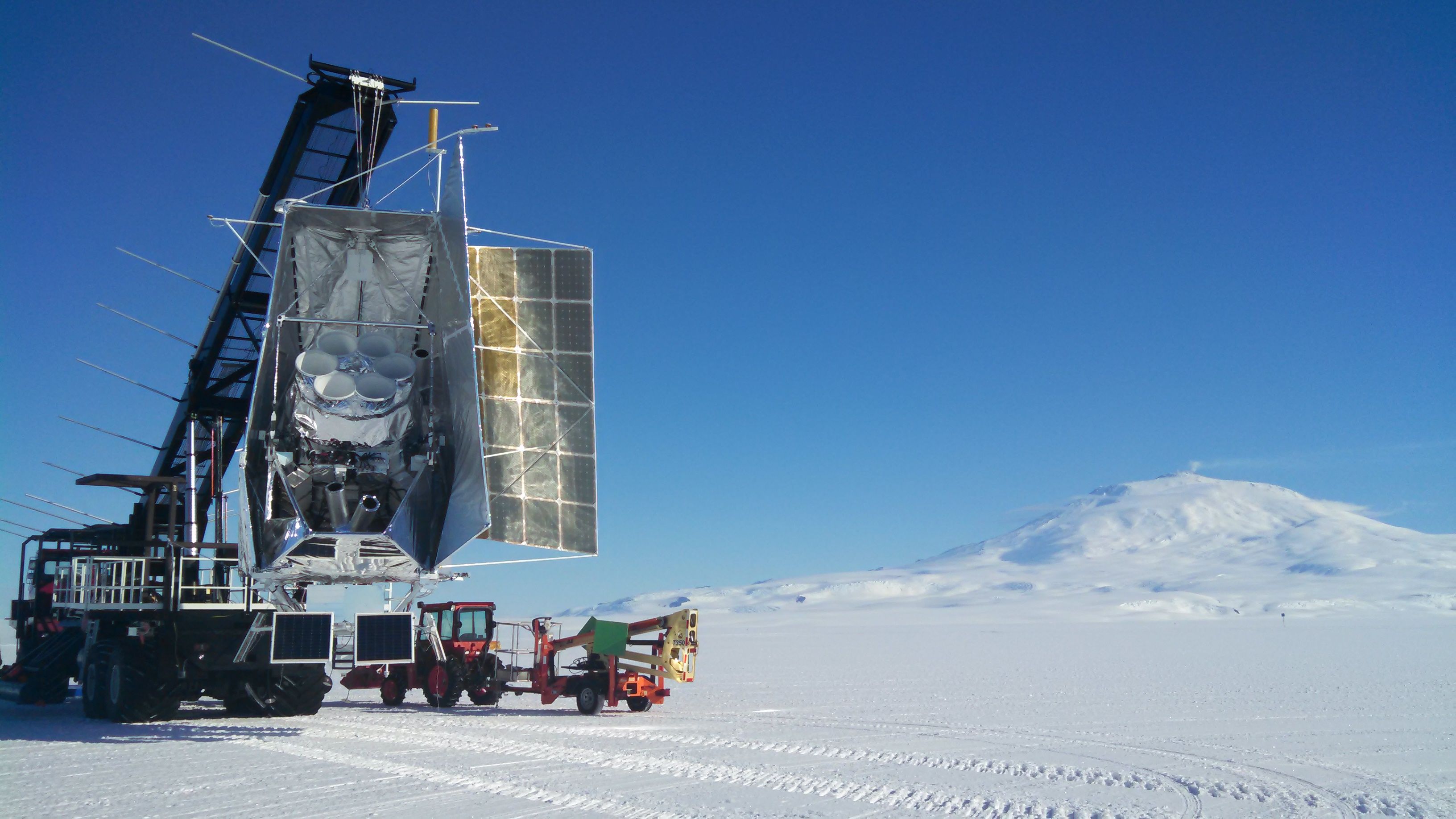 A metallic structure hangs from a wheeled crane in a polar landscape. 