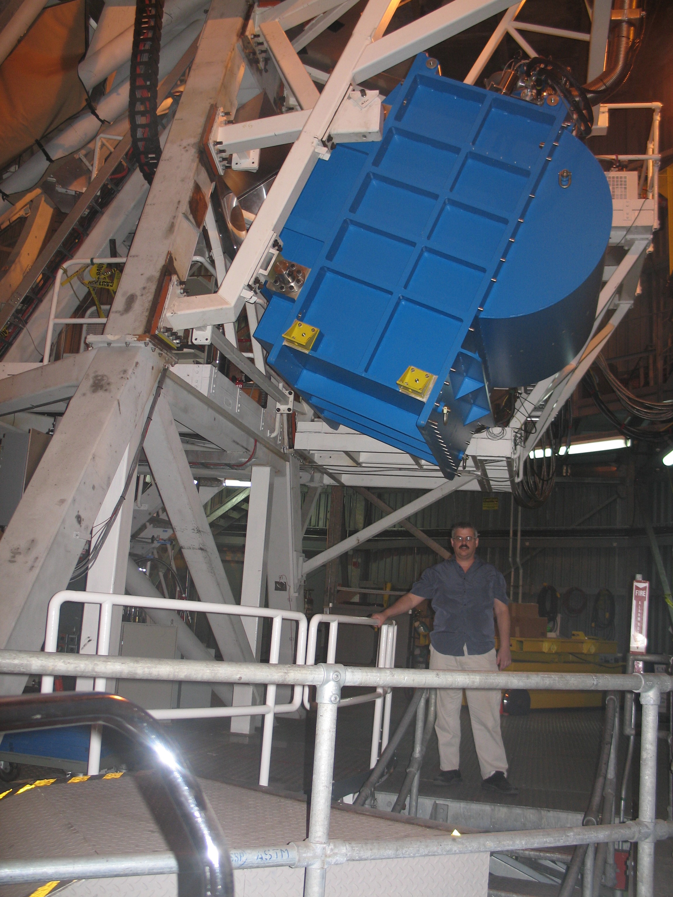 A man stands under a large piece of blue equipment on white scaffolding. 