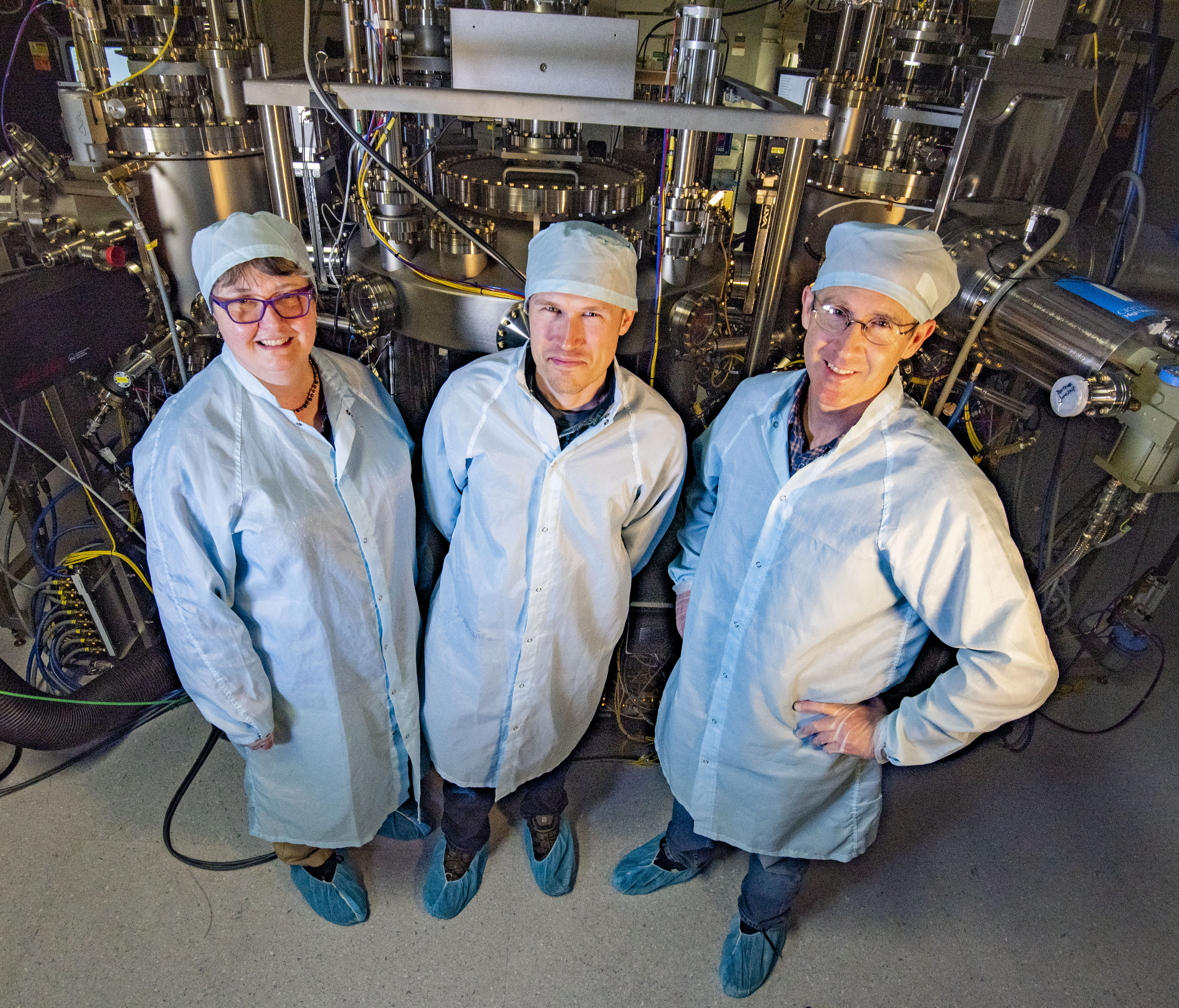 Three people in protective coats and caps pose in front of a large machine with circular parts and connecting hoses.